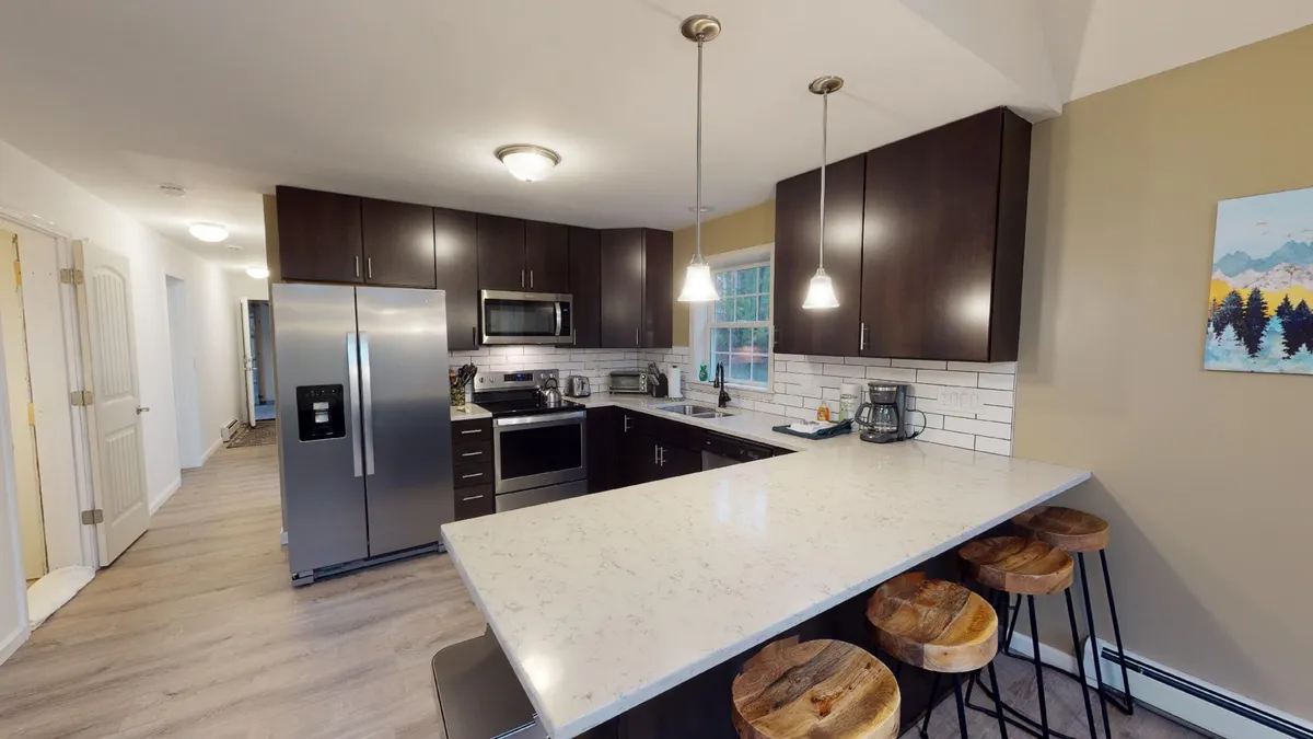 The image showcases a bright, contemporary kitchen with a spacious island and stylish stools, gleaming stainless steel appliances, crisp white countertops, striking pendant lighting, and vibrant wall art. The space feels open and inviting, perfectly set up for cooking, entertaining, and gathering with friends or family.