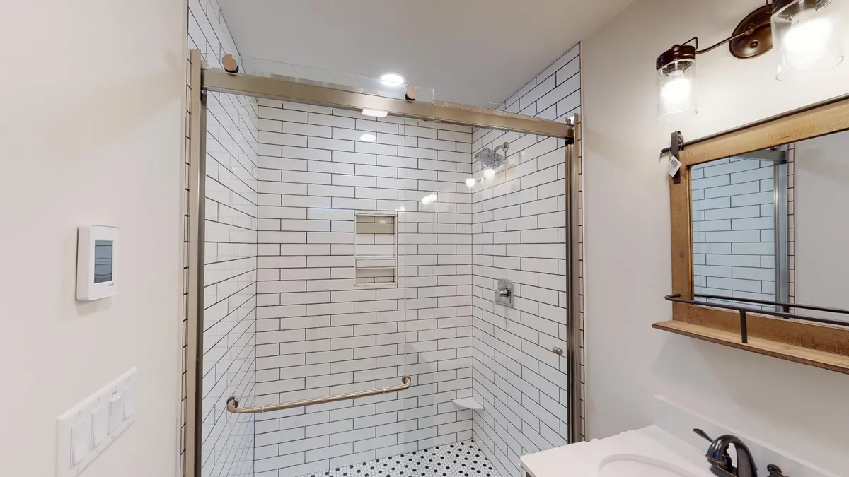 A spa-inspired bathroom boasting a glass shower enclosure, striking black and white tiled floor, wood-framed mirror, and elegant light fixtures. The scene combines modern style with a sense of relaxation.