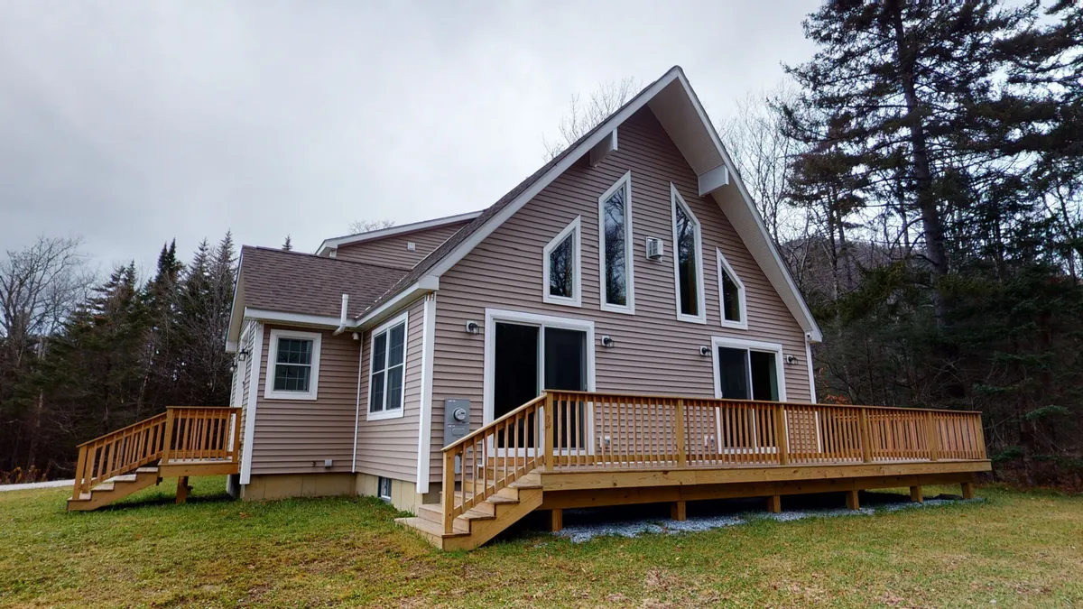 A wide view of a classic wooden wrap-around deck attached to the house, featuring large windows, a charming gabled roof, and surrounded by lush Vermont trees. The space is set for relaxation or gatherings, with plenty of room for outdoor seating and activities.