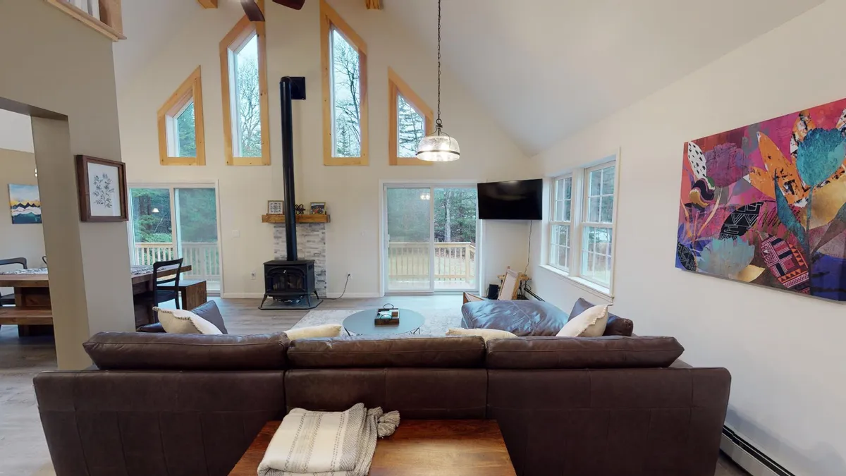 A welcoming main living area with a sofa, artwork, and a dining table framed by expansive windows that let in abundant natural light. The wood stove and cozy furniture create an inviting, comfortable atmosphere, while the view outside suggests a close relationship between the interior and the surrounding natural scenery.