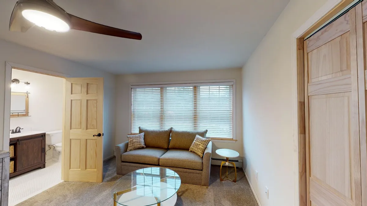 A cozy living area featuring a plush sofa and coffee table next to large windows, allowing plenty of natural light to pour in. The setup creates a welcoming spot for relaxing, enjoying a morning coffee, or taking work breaks. The door to the bathroom is visible, adding to the sense of functionality and flow.