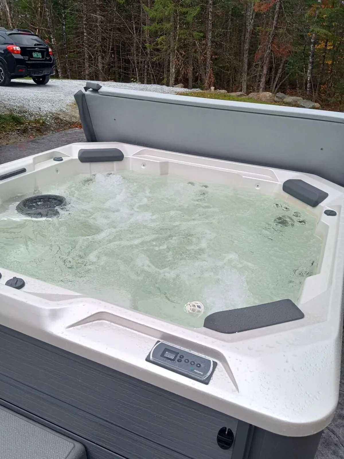 A serene view showing the hot tub in a forested exterior setting, with a gravel driveway in the background. The ambiance is calming, emphasizing the connection between the hot tub and the surrounding nature.
