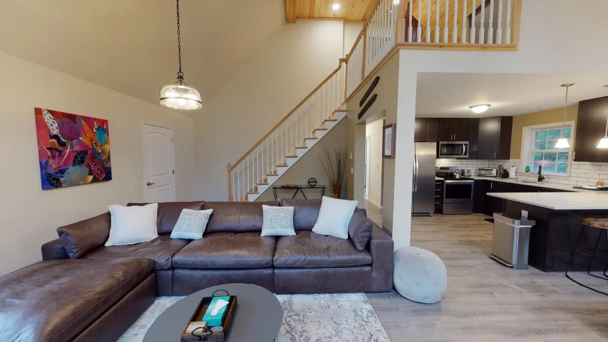 A cozy, inviting living space with a large sectional sofa facing toward a colorful wall art, adjacent to a bright kitchen area and a staircase. The room is spacious and arranged for group gatherings, with plenty of seating to facilitate conversation or movie nights.