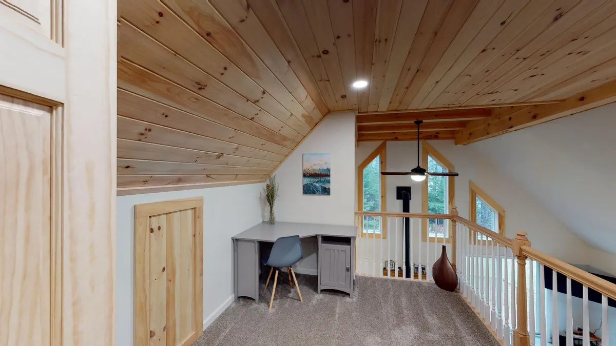 The image features a tranquil loft space set beneath a wooden ceiling, showcasing a desk and chair positioned for productive work or study. Large windows flood the area with natural light, while stylish artwork and a light fixture add character. The view through the railing hints at the open connection to the main living area below, reinforcing the loft’s elevated perspective and serene ambiance.