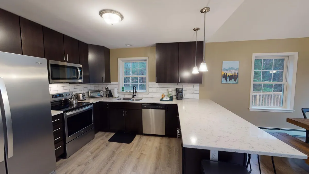 A bright, modern kitchen featuring stainless steel appliances, a marble countertop, an island with seating, and cabinetry bathed in natural light from a window. The space feels both functional and welcoming, ideal for group meal preparation and casual mornings.
