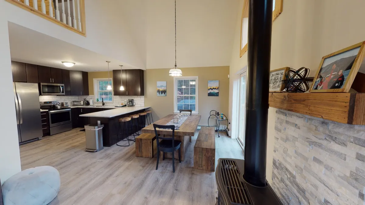 A wide interior shot featuring a large dining table set for a meal, modern kitchen appliances in the background, a stone fireplace, and expansive windows letting in natural Vermont light. The space is open-concept, emphasizing seamless flow between living, dining, and kitchen areas.
