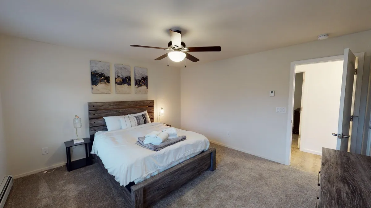 A peaceful bedroom with a neatly made bed, nightstand, wall art, and a doorway leading to private space. The overall mood is calm, highlighting the room's role as a personal sanctuary.