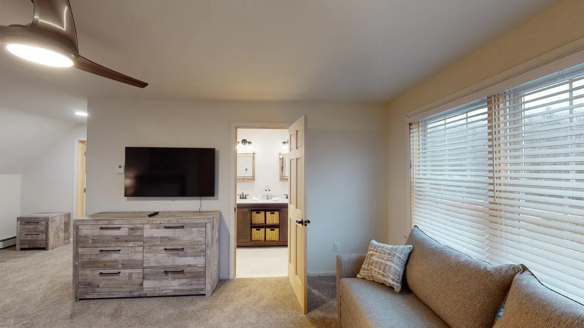 A comfortable living room setup with a sofa facing a TV and a dresser, and a doorway connecting to the bathroom. The windows let in morning sunlight, making it an ideal space for lounging or informal work sessions. The arrangement invites both socializing and quiet moments.